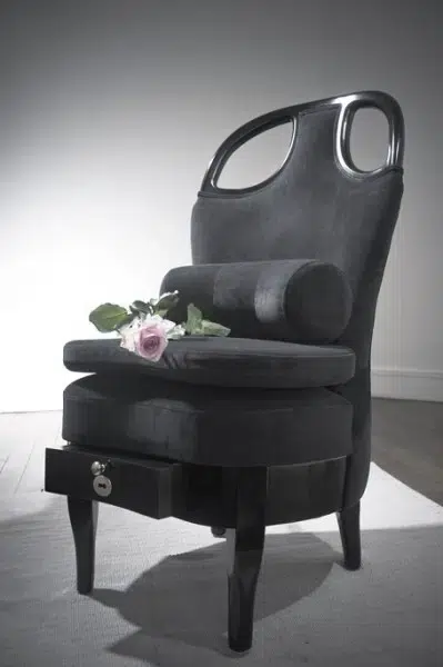 Le fauteuil courtisane, avec du bois laqué vernis, deux poignées sur le dossier, un tiroir intégré, des coussins moelleux et un tissu gris effet velours