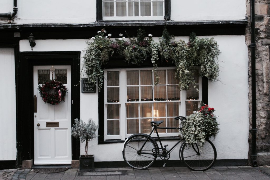 Façade blanche avec large ouverture sans volets, maison anglaise avec un vélo et des fleurs à l'extèrieur