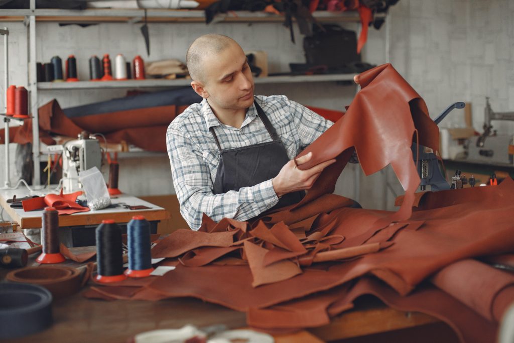 Un artisan examine une pièce de cuir entre ses mains, sur son établi trônent d'autres peaux de mouton