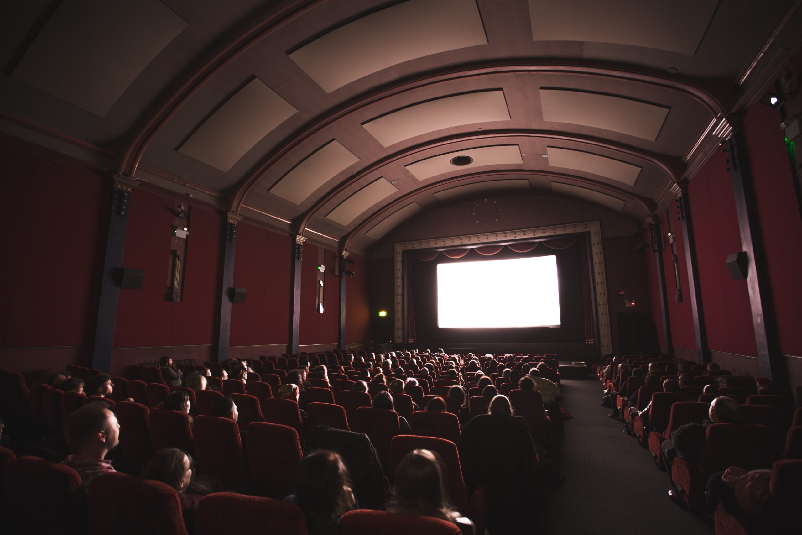 Une salle de cinéma plongée dans la pénombre avec des spectateurs installés sur des fauteuils rouge, observant le grand écran allumé devant eux