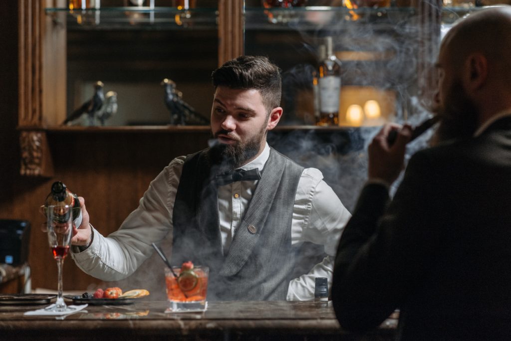 Un serveur en noeud papillon sert un verre à un homme au bar qui fume un cigare, lieu classe haut de gamme